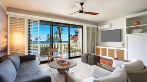 Un vaste espace de vie avec une grande porte-fenêtre coulissante donnant sur un balcon offrant une vue sur les palmiers et l’océan. On y trouve un ventilateur de plafond, un téléviseur à écran plat fixé au mur, ainsi que divers sièges, notamment un canapé, des fauteuils et une table basse ronde.