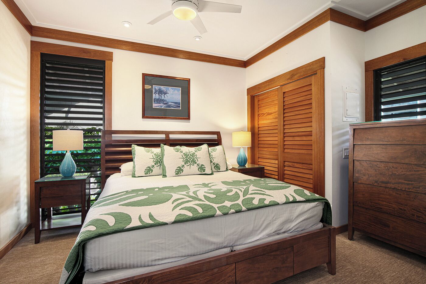 A bedroom with a bed, two bedside tables, a ceiling fan, a framed picture on the wall, and wooden shutters.