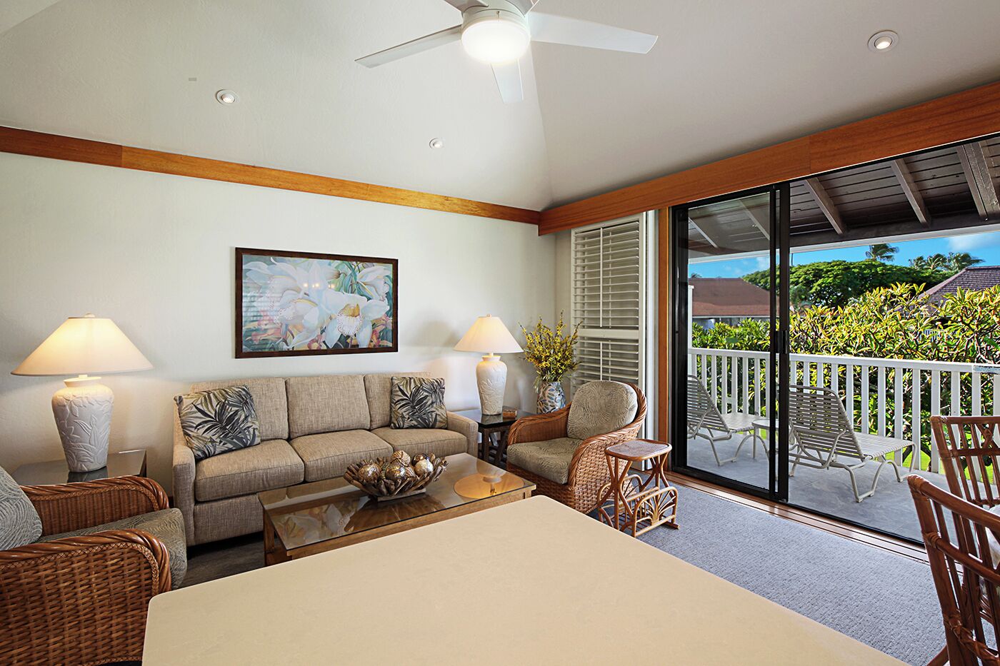A spacious living room with a sofa, two chairs, a coffee table, and a dining table. There is a ceiling fan, a painting on the wall, and a view of the outdoors through a sliding glass door.
