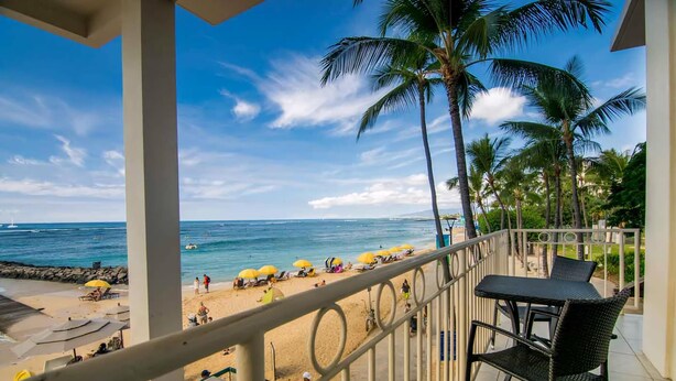 A balcony with a view of a beach, palm trees, and a clear sky. - Waikiki Shore 202 Oceanfront 2 Bedroom (Honolulu)
