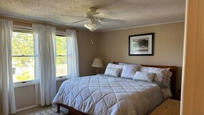 Une chambre à coucher avec un lit, un ventilateur de plafond, une fenêtre avec des rideaux et un tableau encadré au mur.