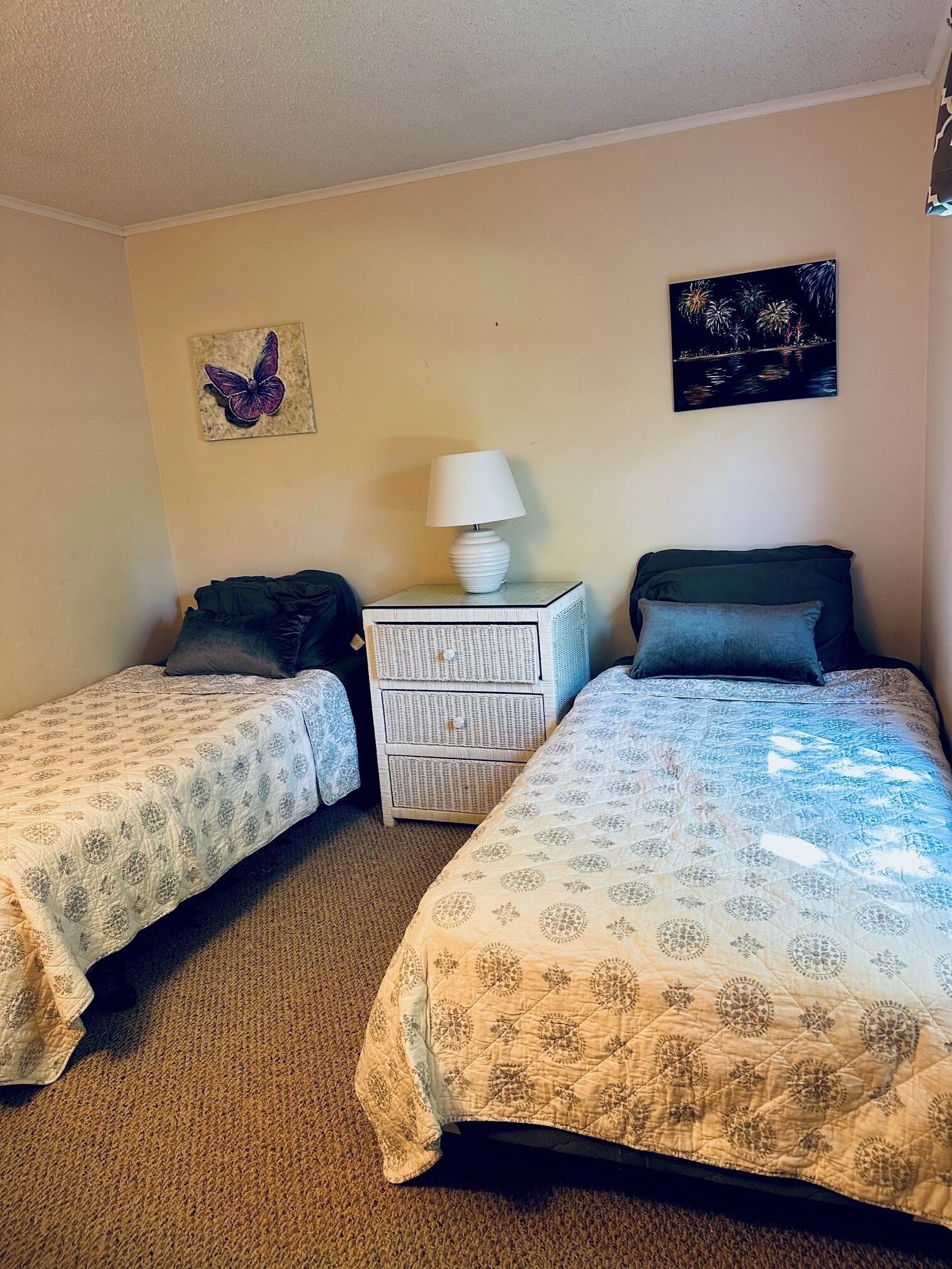 A room with two beds, a nightstand with a lamp, a decorative butterfly wall art, and a night sky painting.