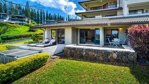 Une maison moderne à deux étages, dotée d’un mur de soutènement en pierre, d’un patio couvert avec mobilier d’extérieur et offrant une vue sur le paysage environnant.