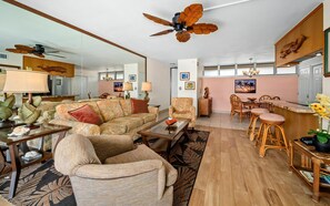 A spacious living area with a sofa, armchairs, a coffee table, a dining area with a bar, and a ceiling fan.