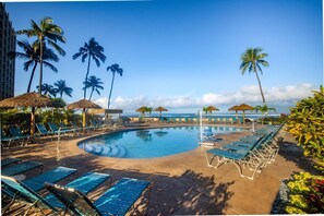 A pool area with lounge chairs and umbrellas, surrounded by palm trees and a clear sky.