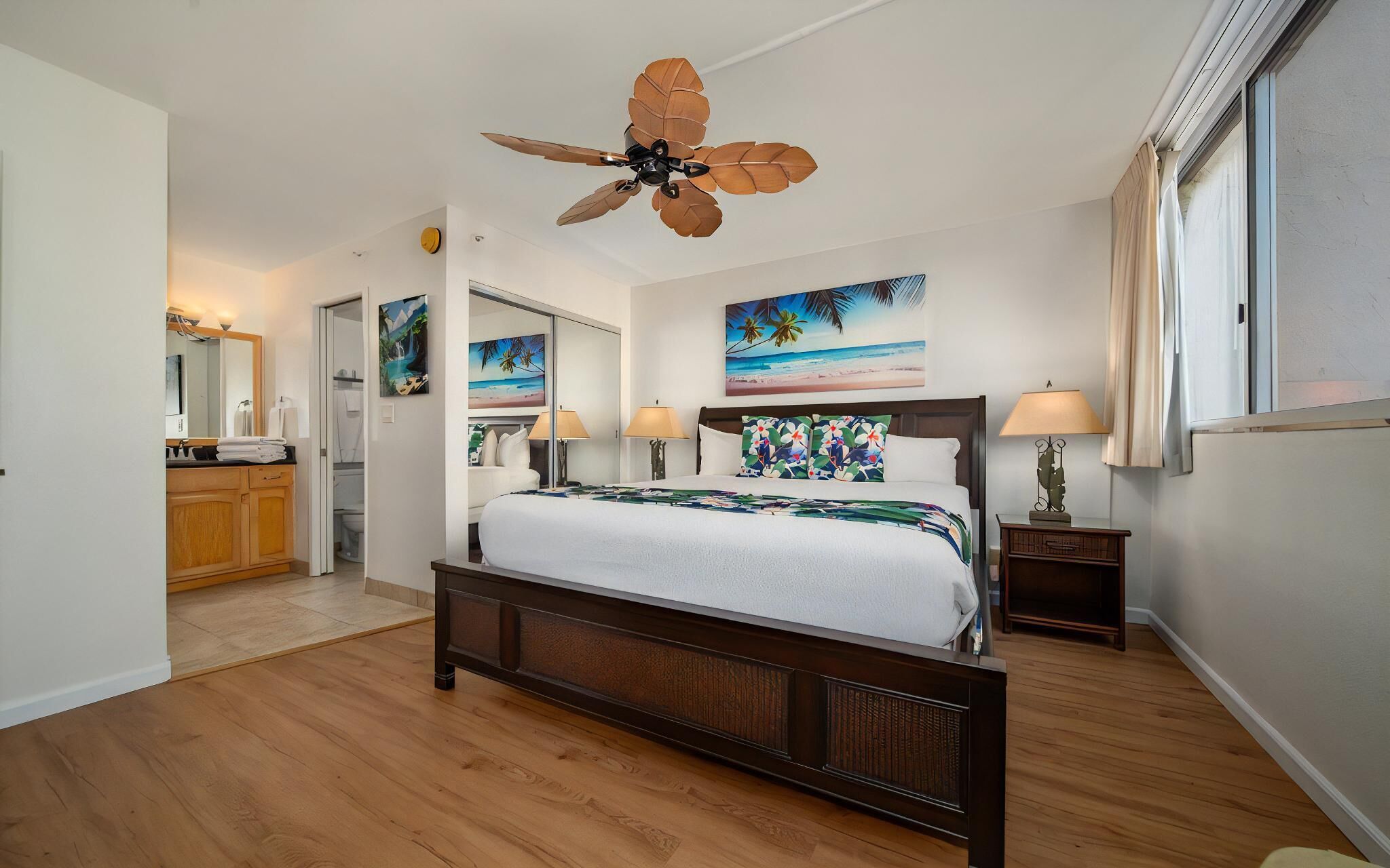 A bedroom with a wooden floor, a bed with a floral bedspread, a ceiling fan, and artwork on the walls.