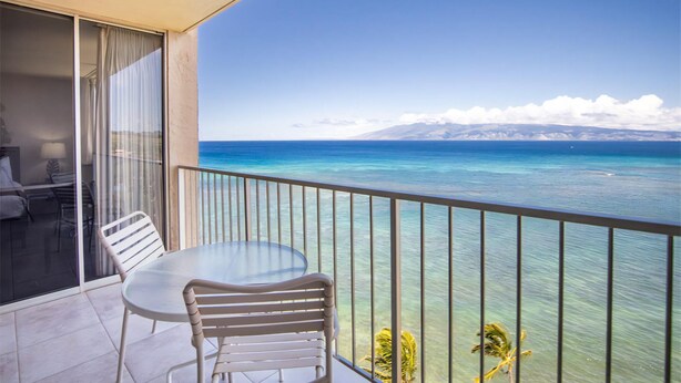 A balcony with a table and chairs, overlooking a clear blue sea and distant mountains.