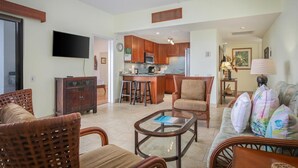 A spacious living room with wicker furniture, a glass coffee table, and a kitchen area with wooden cabinets and a bar stool.