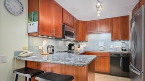 A kitchen with wooden cabinets, a microwave, a refrigerator, a coffee maker, and a dishwasher. There are two bar stools and a clock on the wall.