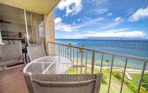 A balcony with a table and chairs, overlooking a beach and ocean with boats.