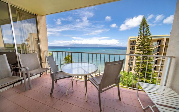 A balcony with a round table and chairs, overlooking a clear blue sea and a distant mountain.