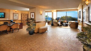 A spacious lobby with a high ceiling, a reception desk, comfortable seating, and large windows offering a view of the ocean.