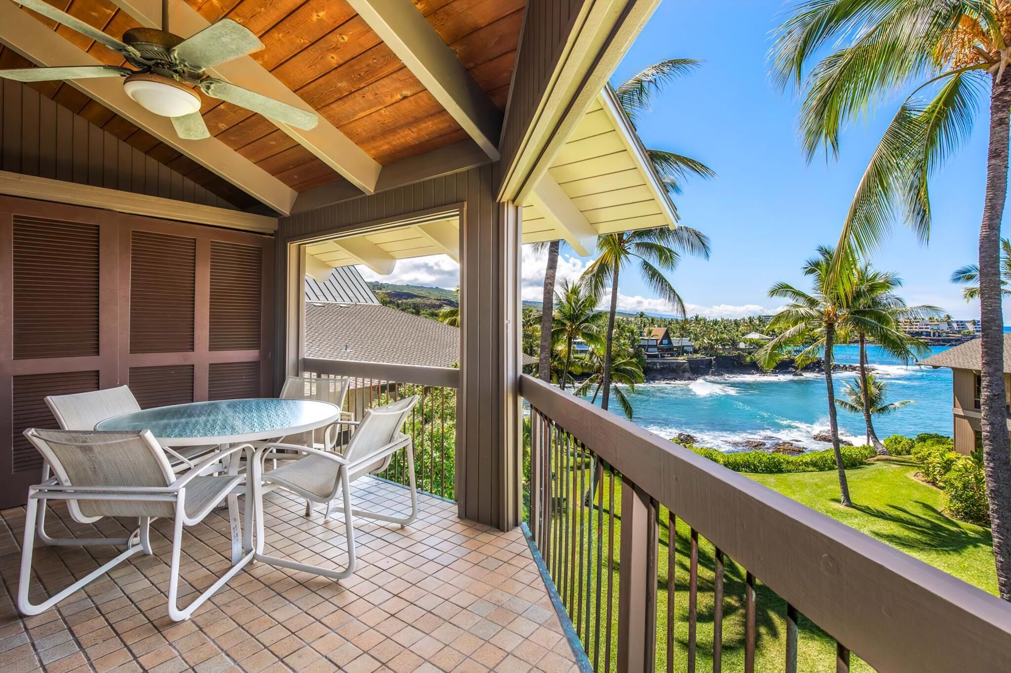 A covered outdoor area with a table and chairs, overlooking a coastal view with palm trees and a clear sky.