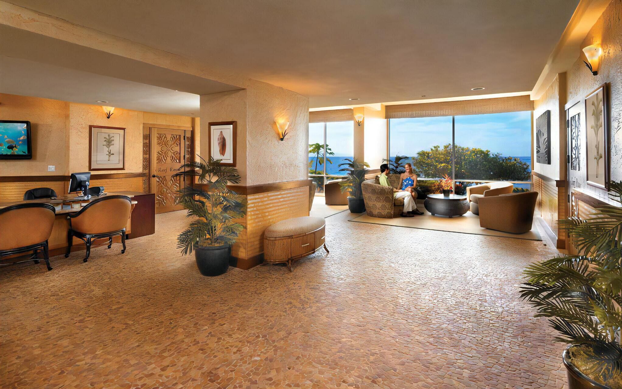 A spacious lobby with a high ceiling, a reception desk, comfortable seating, and large windows offering a view of the ocean.