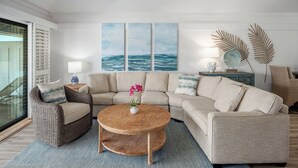 Una sala de estar con un sofá modular, una mesa de centro redonda, un sillón de mimbre y vistas al océano a través de amplios ventanales.