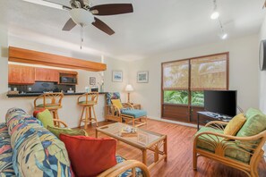 Una sala de estar con sofá, sillas, una mesa de centro, televisor y una zona de cocina con microondas y armarios.