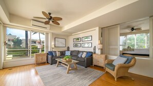 A spacious living room with a sofa, coffee table, and wicker chair, large windows with blinds, ceiling fans, and a view of buildings outside. - Palms at Wailea Villa 806 Ocean View 2BD 2BA (Wailea)