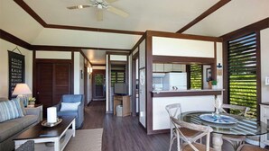A spacious living area with a sofa, coffee table, and a glass dining table with chairs. There is a kitchen area with a refrigerator and a window with blinds.