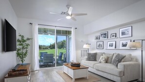Un salón con sofá, mesa de centro, televisor, ventilador de techo y vistas al exterior.