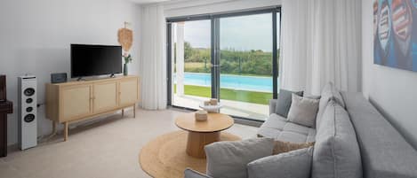 A modern living room with a grey sofa, a wooden cabinet with a flat-screen TV, a round coffee table, and a large window overlooking a pool.
