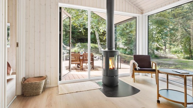 A cozy room with a wood-burning stove, wooden flooring, a large sliding glass door leading to an outdoor deck, and a view of a lush garden. - 3 bedroom amazing home in Nykøbing Sj (Nykøbing Sj)