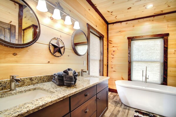 Baño de cabaña de madera con lavamanos de encimera de granito, dos espejos, una bañera blanca y una ventana con persianas.