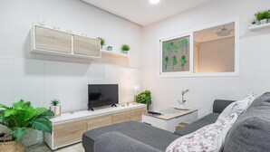Un salon moderne avec un canapé gris, un téléviseur à écran plat, un meuble en bois et une petite table d’appoint sur laquelle se trouvent une lampe et une plante.