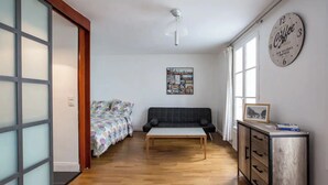 A compact living space with a wooden floor, a sofa, a bed with colorful bedding, a small table, and a wall clock. - Well-appointed St. Michel studio (Paris)