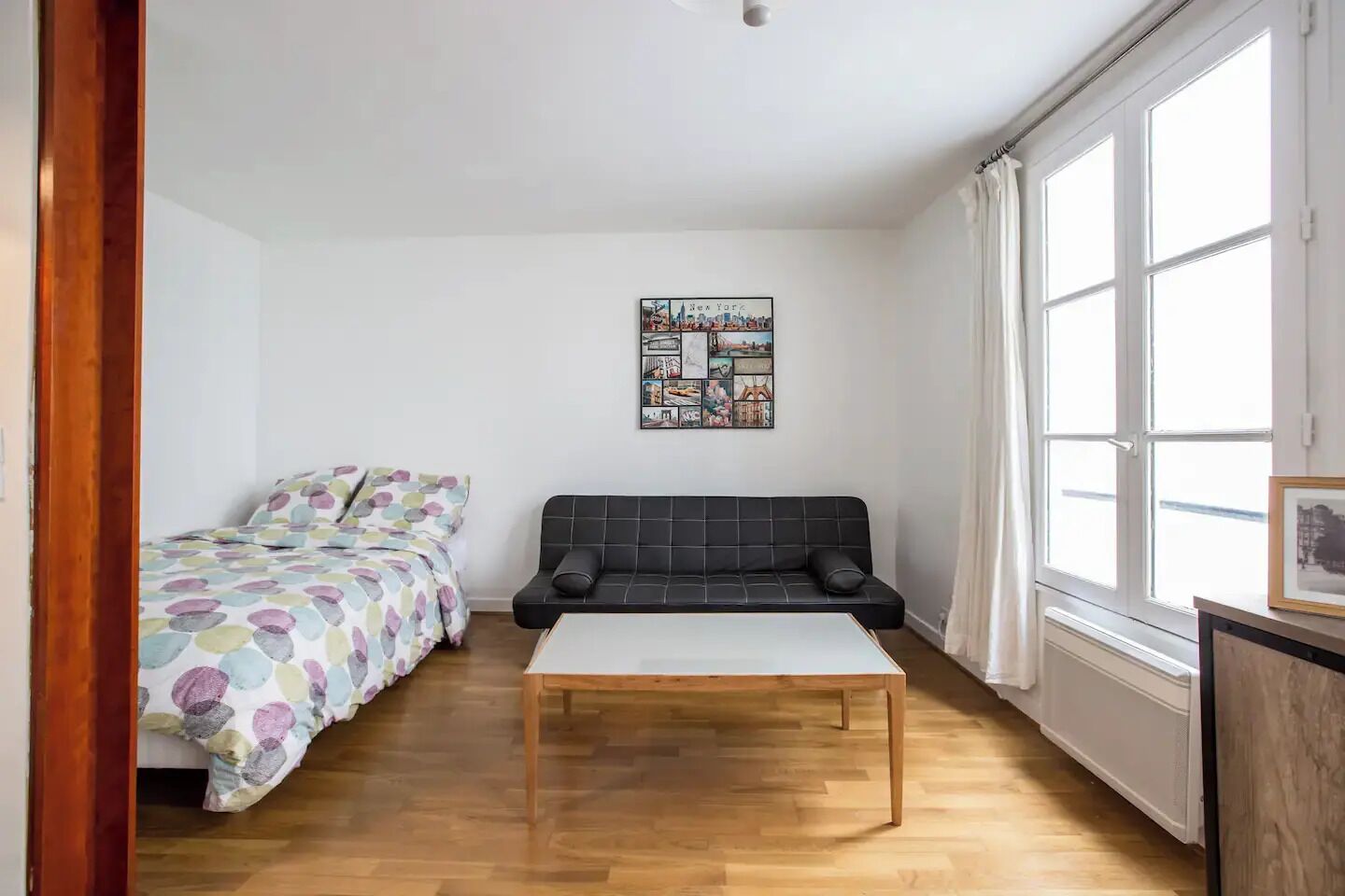 Une petite pièce lumineuse, avec un lit, un canapé et une table. Un grand mur-fenêtre se trouve d’un côté, et l’autre mur est décoré d’une œuvre d’art représentant un paysage urbain.