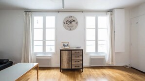 A spacious room with wooden flooring, a white table, a cabinet with drawers, and a wall clock. - Well-appointed St. Michel studio (Paris)