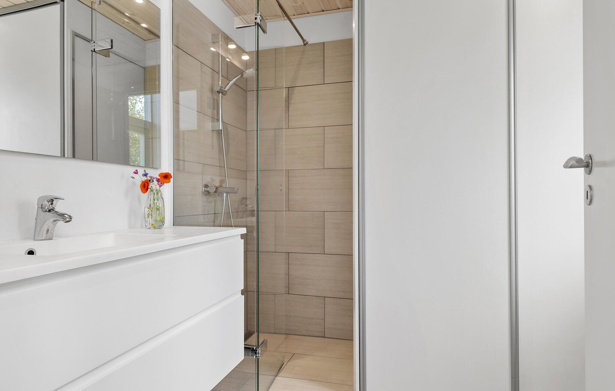 A modern bathroom with a glass shower enclosure, a white vanity with a mirror, and a vase with flowers.