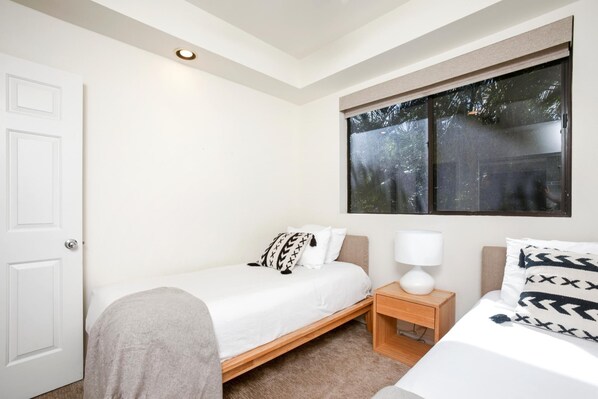 A bedroom with a single bed, a bedside table with a lamp, a window with a view of trees, and a door.