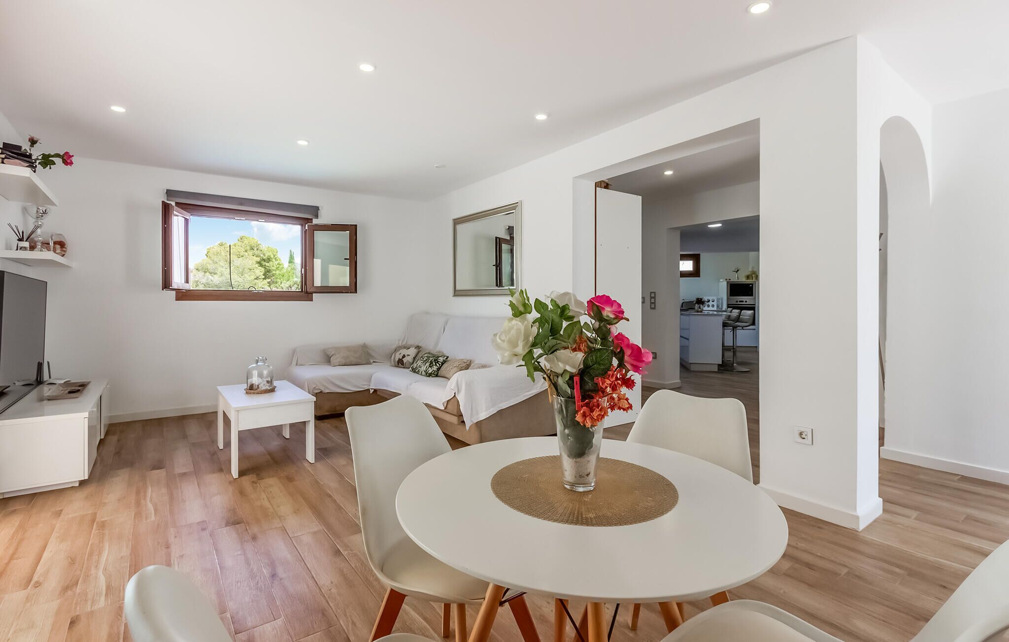 Ein modernes Wohnzimmer mit Essbereich, einer Sofagarnitur, einem Couchtisch mit Blumenvase und Blick zur Küche durch einen offenen Durchgang.