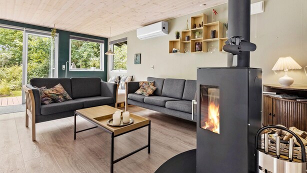A modern living room with a wood-burning stove, grey sofas, a wooden coffee table, and a view of greenery through large windows. - 2 bedroom nice home in Rønde (Rønde)