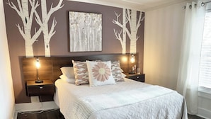 Un dormitorio con una cama grande, cabecera de madera, dos mesitas de noche con lámparas, un cuadro colgado en la pared y una ventana con cortinas.