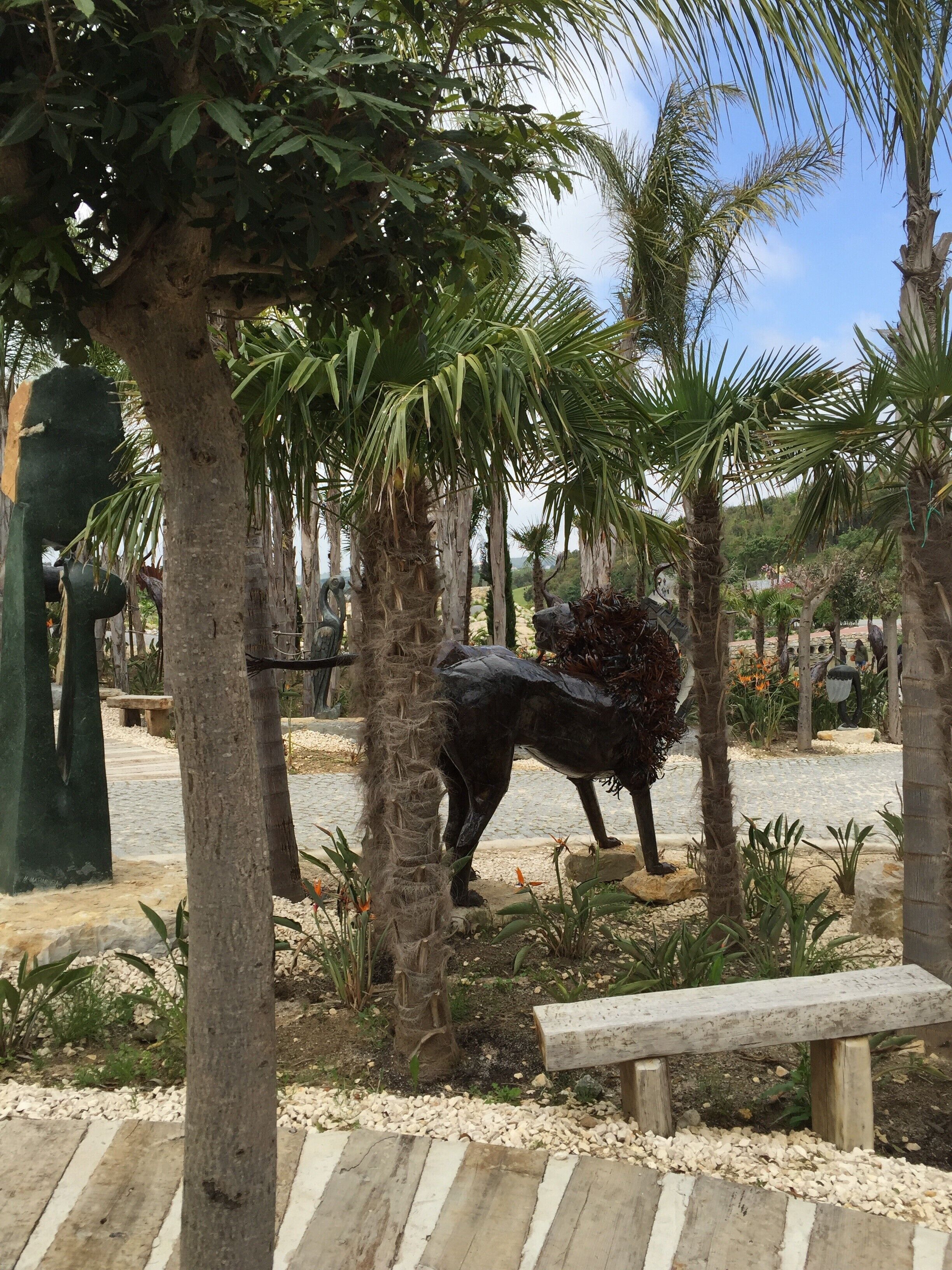 Een beeld van een paard in een tuin met palmbomen en een stenen bank.