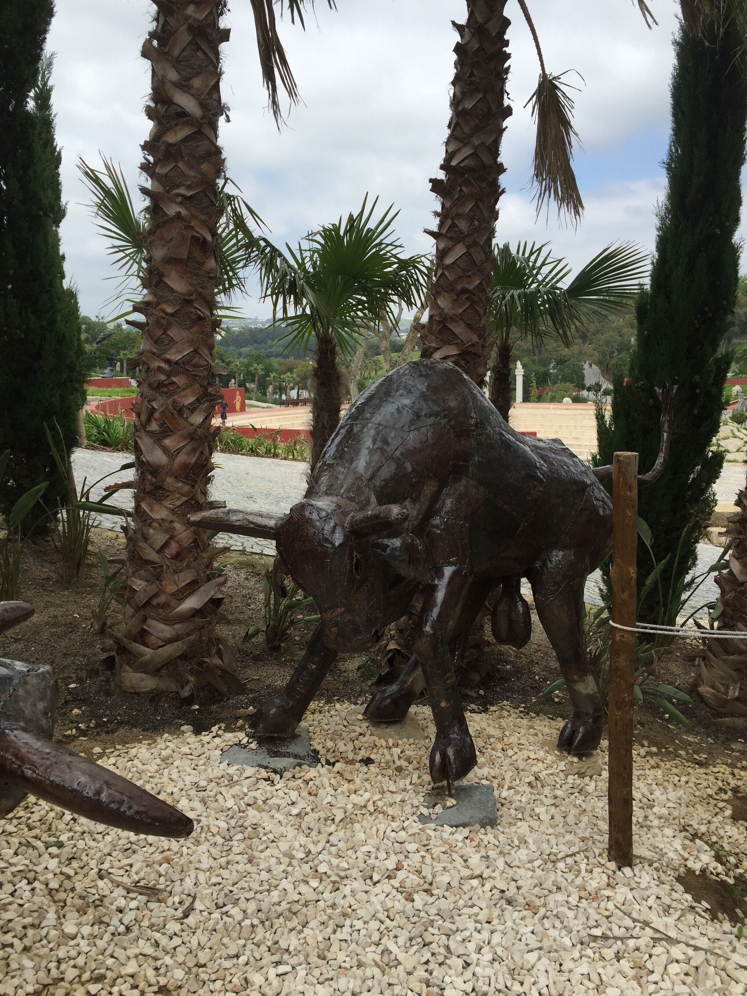 Estátua de bronze de um touro em um jardim com palmeiras e um caminho de cascalho.