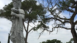 En stenskulptur som föreställer en figur med en krona och flera ansikten, omgiven av träd och en vattenyta.