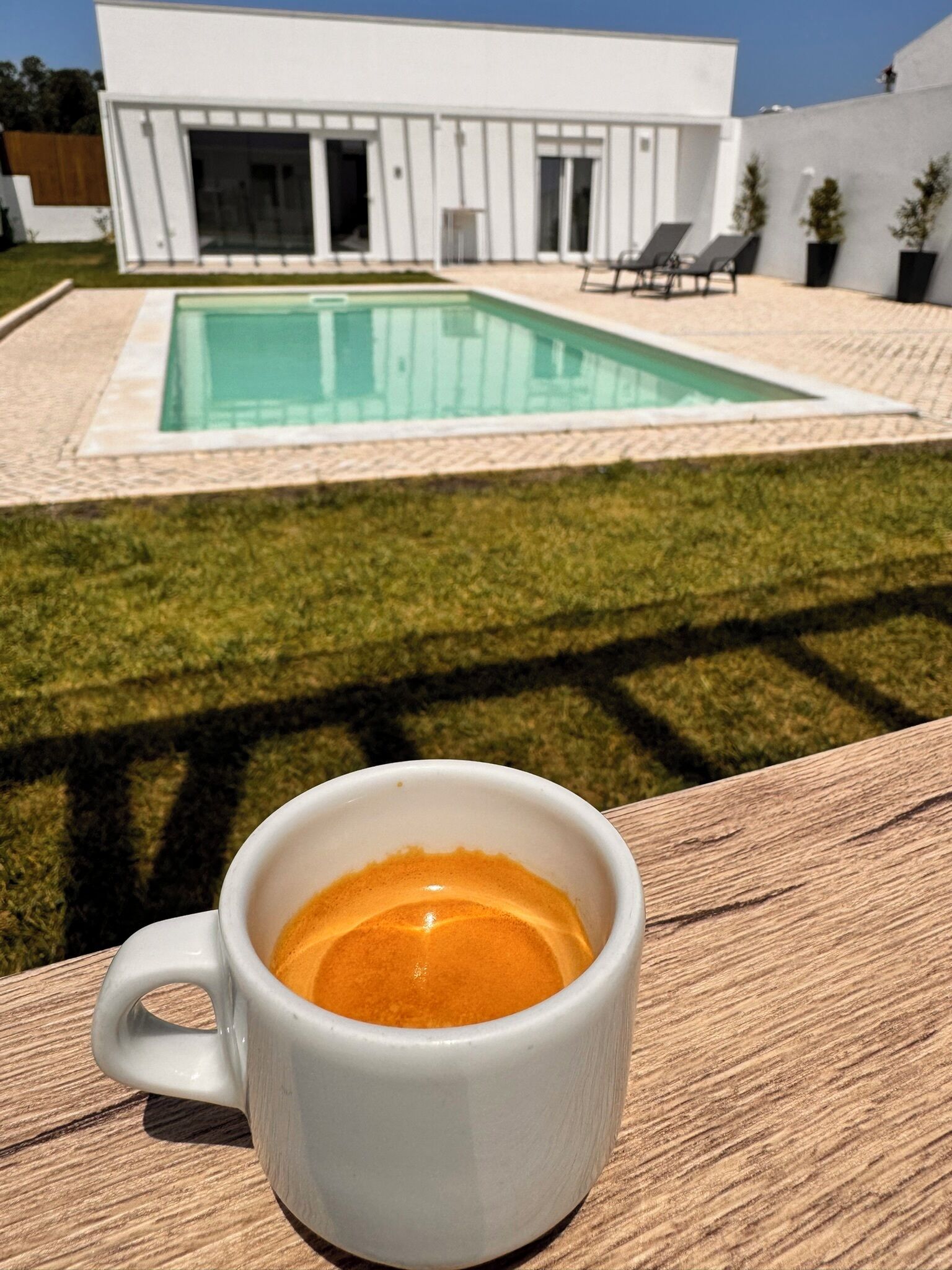A white mug with a beverage on a wooden surface. In the background, there is a modern white building with a swimming pool and a well-maintained lawn.