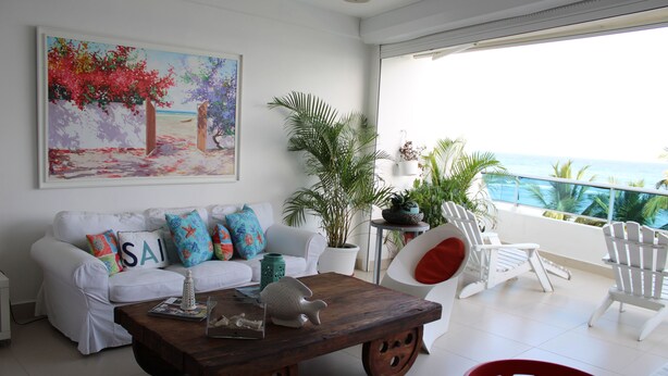 A living room with a white sofa, colorful pillows, a wooden coffee table, and a painting of a beach scene. - Las Olas 2BR 6PAX 3Bds HugeTerrace (Guayacanes)