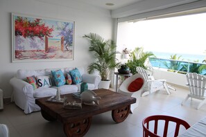 A living room with a white sofa, colorful pillows, a wooden coffee table, and a painting of a beach scene. - Las Olas 2BR 6PAX 3Bds HugeTerrace (Guayacanes)
