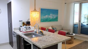A modern kitchen with a granite countertop, stainless steel sink, and a dishwasher. There is a dining area with a red chair and a round table. A painting of the ocean is on the wall, and a large window provides a view of the outdoors. - Las Olas 2BR 6PAX 3Bds HugeTerrace (Guayacanes)