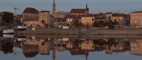 A riverside cityscape with historic buildings, a prominent church tower, and a calm water body reflecting the architecture.