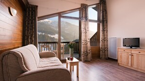 A cozy room with a sofa, a small table with a vase and candles, a TV, and a view of mountains through large windows. - Appartement à Hauteluce, navette gratuite (Hauteluce)