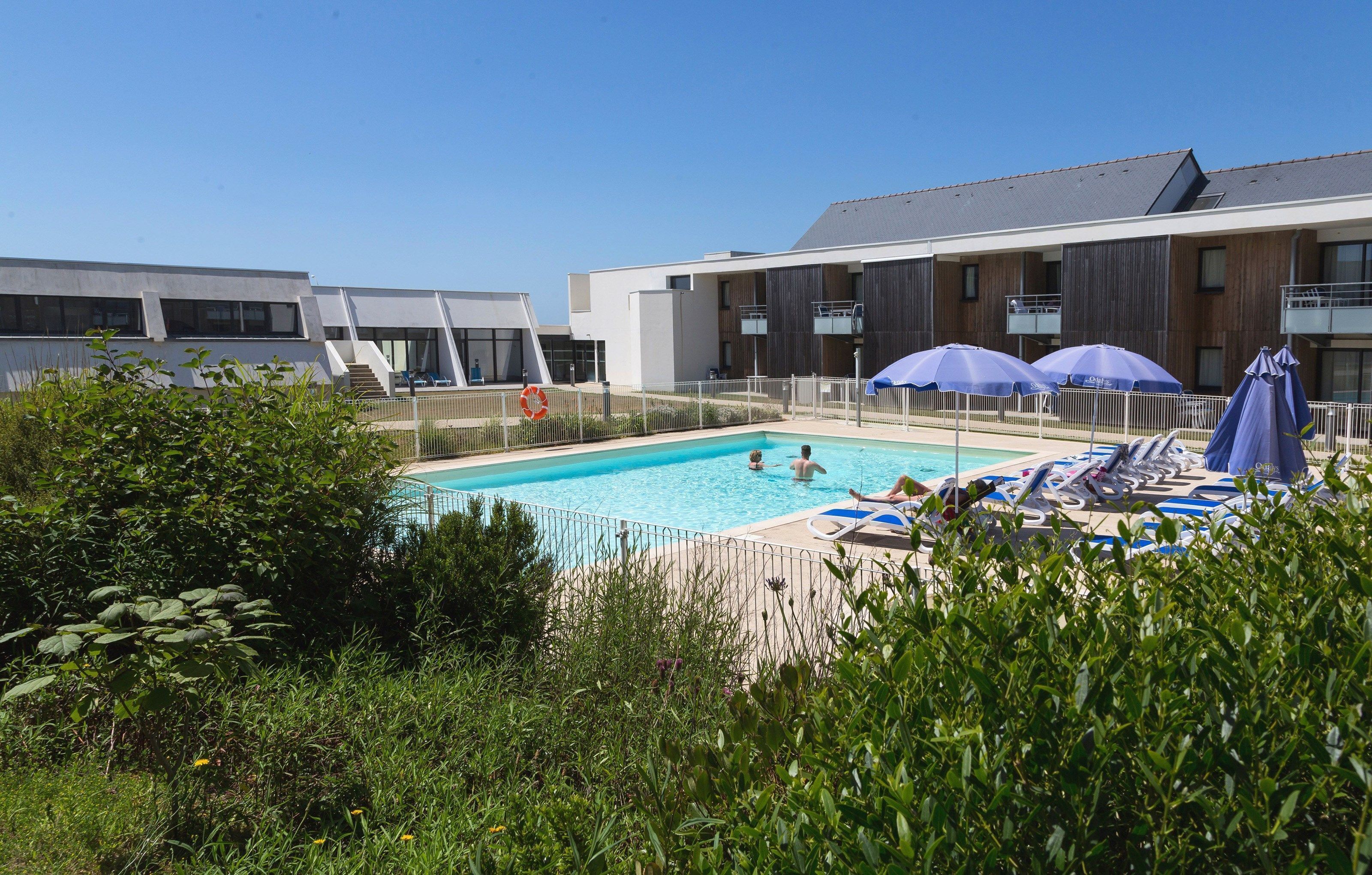 Un espace piscine doté de chaises longues et de parasols, entouré de verdure et de bâtiments modernes.