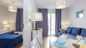 A hotel room with a bed, a sofa, a dining table with breakfast items, and a desk with a lamp. - Appart cosy et équipé sur le lac | Wi-Fi Gratuit ! (Hourtin)