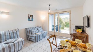 Un salon confortable avec un canapé, une table à manger dressée avec du pain et des fruits, et un téléviseur fixé au mur.