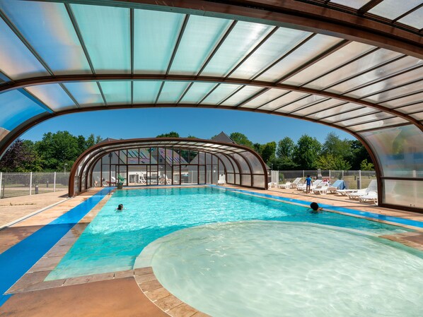 A covered swimming pool with clear blue water, a curved roof, and a view of a house in the background.