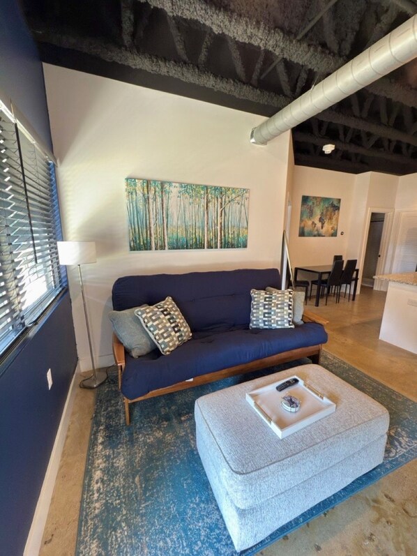 Uma sala de estar moderna com um sofá azul escuro, almofadas estampadas, um banquinho cinza e uma pintura de uma floresta.