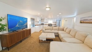 Ein modernes Wohnzimmer mit einem großen Flachbildfernseher, einem beigefarbenen Ecksofa, einer Holzkonsole und einem kleinen Hocker.